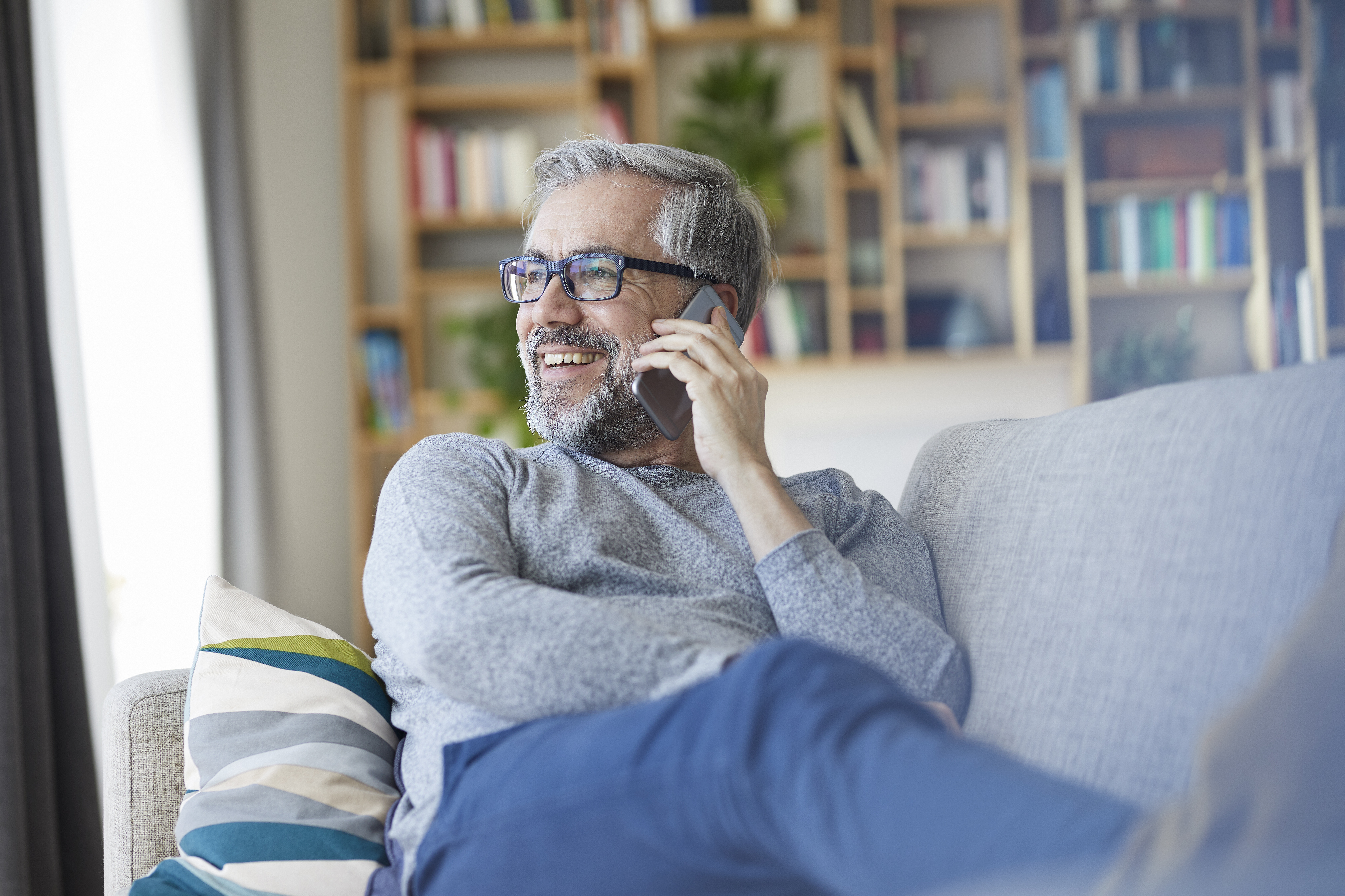 Mature man on the phone sitting on couch at home looking out of window