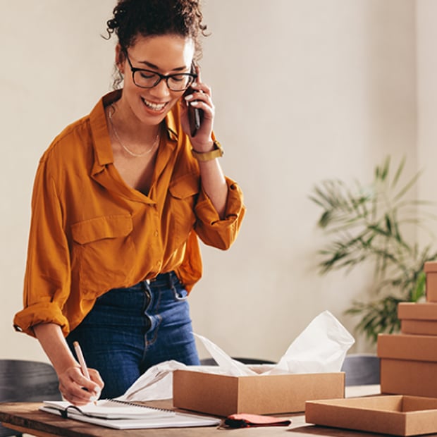 What Is VoIP Phone System Thumbnail Photo of a smiling woman standing at her busy, cluttered desk in what appears to be a home office. She is holding her cell phone, listening and taking notes.